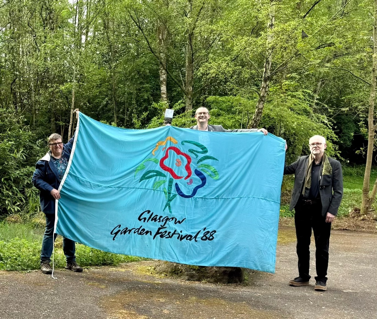 Kenny Brophy, Gordon Barr and Lex Lamb, holding a large blue Glasgow Garden Festival ’88 flag in Festival Park.