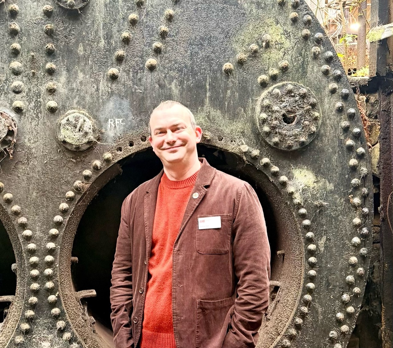 Gordon Barr standing in front of a large, weathered riveted metal boiler in a mining museum.