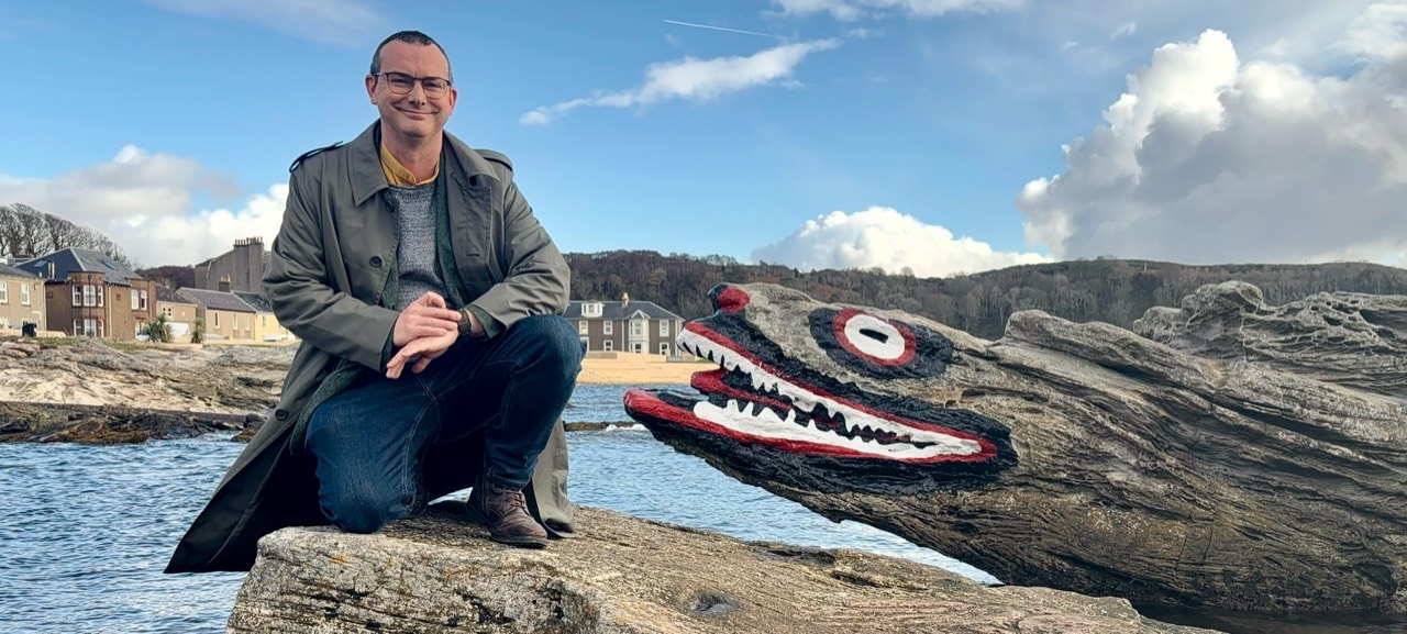 Gordon Barr crouching beside Crocodile Rock in Millport - a seaside rock painted to resemble a crocodile’s head.