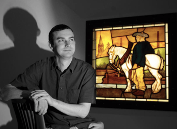 Gordon Barr seated in Maryhill Burgh Halls, beside a 1878 stained-glass panel depicting a man with a white horse next to the Forth & Clyde Canal.